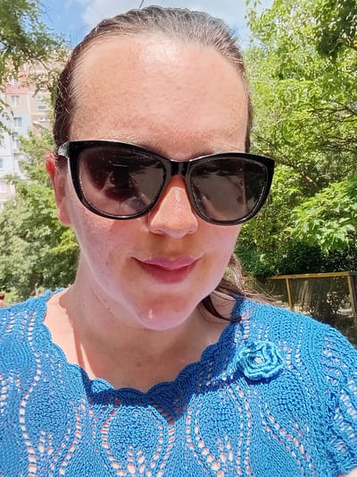 A medium shot portrait shows a woman wearing black sunglasses and a blue knitted top with a floral embellishment. She is looking slightly off-camera, with her head tilted. The background consists of green trees and a building, suggesting an outdoor urban setting. The lighting indicates it is daytime. There is no evidence to place this image in Kherson, Ukraine.