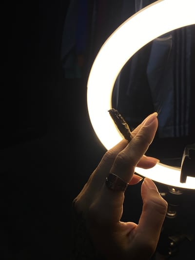 A close-up shot focuses on a person's hand holding a partially smoked, dark-colored cigarette or joint. The hand has a gold ring on one of the fingers and a tattoo visible on the wrist. The background features a circular light ring that provides illumination, with the rest of the scene mostly dark. The setting suggests an indoor environment with a modern light source. There is no evidence that places the scene in Llavallol, Argentina.
