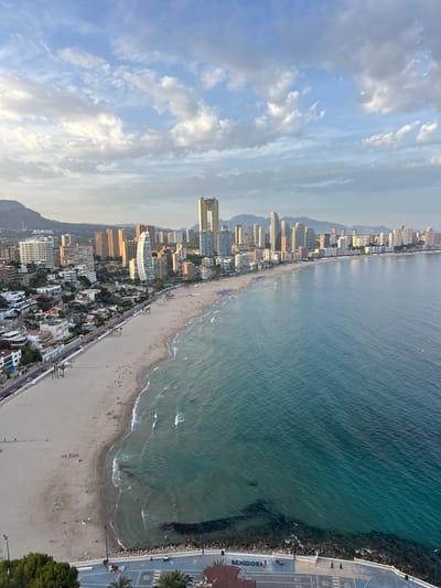 The image shows a wide-angle, aerial view of a beach and cityscape in Benidorm, Spain. The foreground features the turquoise waters of the Mediterranean Sea meeting a sandy beach dotted with sunbathers and loungers. Waves gently lap the shore. Along the beach, a promenade runs parallel, lined with trees, streetlights, and some pedestrians. Further inland, a densely populated urban landscape unfolds, characterized by numerous high-rise buildings of varying architectural styles and heights, stretching along the coastline. Some of these buildings are hotels or apartment complexes. In the background, mountains form a natural backdrop to the city. The sky is partially cloudy, with soft sunlight casting a warm glow over the scene. The overall impression is of a vibrant coastal resort town.