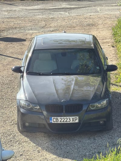 A grey BMW sedan is parked on a gravel surface next to a dirt road with green vegetation on one side. The car's license plate reads "CB 2223 XP" with the country code "BG" for Bulgaria. No people are visible. The location is estimated to be Petarch, Bulgaria, based on the license plate. The scene depicts a static, outdoor setting.