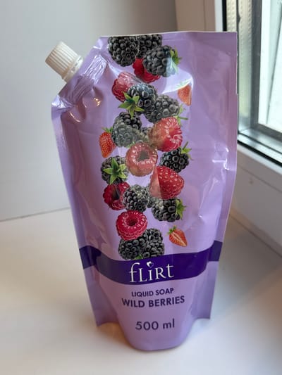 A pouch of liquid soap with a wild berry scent is shown. The pouch is lavender in color and features an illustration of various berries, including raspberries and blackberries. The brand name "FLIRT" is printed in white text, followed by "LIQUID SOAP WILD BERRIES" and "500 ml". The pouch has a white screw-top cap and is positioned on a white surface, possibly a windowsill or table, with a window visible in the background. The lighting suggests it is daytime. The scene is indoors.