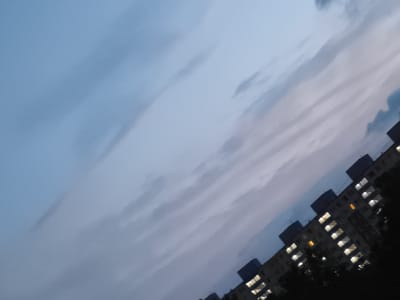 The image is a tilted, low-light photograph showing a section of a residential apartment building at dusk or night. The sky is a muted blue with wispy grey clouds stretching diagonally across the frame. The building, which appears to be multi-story, is silhouetted against the sky with many windows illuminated, indicating that people are inside. The architecture suggests a Soviet-era block, common in Eastern European cities. The angle of the shot and the lighting suggest it was taken from ground level or a lower floor of another building, looking upwards. The city of Most, Czechia, is known for its numerous apartment blocks built during the communist era, often referred to as "panelák" buildings, which this structure resembles.