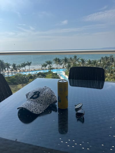 A table on a balcony overlooks an ocean view with palm trees. A chair, a can of hard seltzer, sunglasses, and a baseball cap are on the table. The sky is blue with some clouds.
