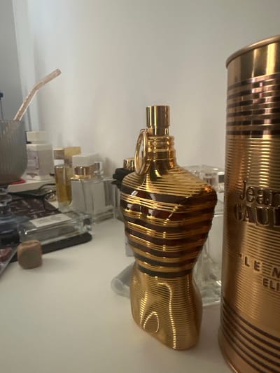 The image displays a collection of perfume bottles arranged on a shelf against a plain, light-colored background. The primary focus is on two distinct perfume bottles, both with a striking gold, ribbed design. The top bottle is cylindrical with vertical gold and dark stripes. The text "jean paul" is visible on this bottle. Below it, a more uniquely shaped bottle, also gold and ribbed, is positioned horizontally. Its form resembles a bust or torso. Other perfume bottles, varying in size and color, are visible in the background, some appearing to be clear glass with gold accents, and others darker. A slender, clear tube-like object is also present in the lower left portion of the frame. The lighting is bright, suggesting an indoor setting, possibly a well-lit room or display area. No people are visible in the image. The overall impression is one of a curated collection of luxury fragrance items. The provided location context of Madara, Bulgaria, does not offer any direct visual cues within the image itself.