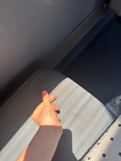 A person's hand is shown in the foreground, making a finger heart gesture. The hand is positioned over a grey, textured seat. Sunlight casts a strong shadow across the seat and the hand. In the background, there is a dark blue area which appears to be another seat or surface. The overall environment suggests the interior of a vehicle, possibly a bus or train. The image does not provide any specific information about the city of Pudong, China.