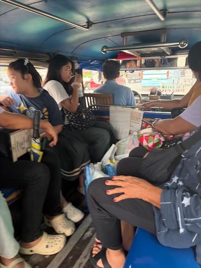 The image shows a group of people packed inside what appears to be a type of public transport, possibly a jeepney, in Dagupan, Philippines. The setting is cramped and appears to be during the daytime, as indicated by the bright lighting. The people are seated closely together, with some standing or leaning against the vehicle's interior.
Visible subjects include several individuals of various ages and genders. One person in the foreground is wearing a dark blue t-shirt with a star pattern and black pants, with their hand resting on their lap. Another person in the middle ground is wearing a light blue shirt and has their head turned away. A woman in the foreground is wearing a white t-shirt and has her head tilted up. There are also glimpses of other passengers, including children, in the background.
Objects present include bags, some of which are colorful and patterned, placed on laps and the floor. There are also what look like white plastic bags, possibly containing purchases or supplies. A metal handrail runs along the side of the vehicle, providing support for passengers. The interior of the vehicle is visible, with blue and green surfaces, and wooden benches or seating.
The activity is transport, with people traveling together in a crowded vehicle. The mood appears to be one of everyday travel, with no overt signs of distress or excitement. The closeness of the passengers suggests a common mode of local transportation where space is at a premium.
Notable details include the footwear of some passengers, with at least two people wearing light-colored Crocs-style clogs. The woman in the foreground has bright red nail polish. The image is taken from a high angle, looking down into the vehicle.
There is some visible text in the background, including what appears to be writing on a sign or advertisement inside the vehicle. Some of this text is in a non-Latin script, possibly Filipino or another regional language. There's also a license plate or vehicle identification number visible through the window which reads "D7E1 637".