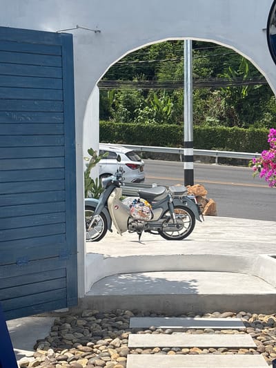 The image, likely taken in Kamala, Thailand, presents an oblique view of an outdoor setting. A blue, vertically-planked gate dominates the lower portion of the frame. Above the gate, a white, semi-circular archway frames the scene. Through the archway, there's a road, a roadside hedge, and a power pole with black and white bands. A scooter is parked partially within the frame of the archway, close to the road. Behind the scooter is a parked white car. To the right of the scooter, outside the arch, is a retaining wall of rounded rocks and what appear to be concrete slabs. A flowering plant with pink blossoms is visible above the road.