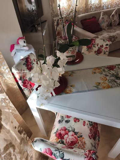 The image depicts an indoor scene, likely a living room or dining area, in Livorno, Italy. A white table is centrally positioned, adorned with a floral runner and a white orchid plant in a pot matching the runner's floral pattern. The table is surrounded by chairs, one of which is partially visible with a floral-patterned seat cushion. To the left, a white cabinet holds a plush unicorn toy with pink accents. In the background, a couch with floral cushions and a dark red decorative pillow is visible, along with patterned curtains covering a window. The lighting suggests it is daytime, with sunlight filtering through the curtains, casting shadows. The overall impression is of a home interior decorated with floral motifs.
