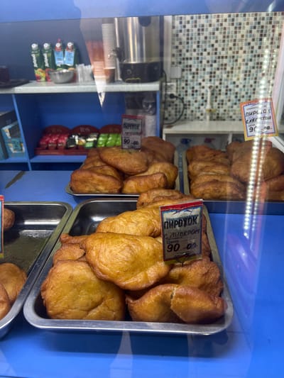 This is a photograph taken inside a store, likely a bakery or a small grocery store, in Lomonosov, Russia. The primary subjects are trays of fried pastries, possibly pirozhki, displayed on a bright blue surface. The pastries are golden brown and appear to be freshly made, with some glistening from oil.

There are several price tags visible on the pastries. One tag, in Russian, reads "ПИРОЖОК С КАПСТОЙ" (Pirozhok with cabbage) and "ЦЕНА 90-00" (Price 90-00). Another tag identifies "ПИРОЖОК С ЛИВЕРОМ" (Pirozhok with liver), also priced at 90-00. The presence of these price tags indicates the pastries are for sale.

In the background, there is a tiled wall and a glass display case. Inside the display case, stacks of what appear to be beverage cartons or juice boxes are visible, along with some disposable cups. Reflections on the glass suggest indoor lighting, possibly artificial, and the overall scene seems to be indoors during the day. The atmosphere appears to be that of a commercial establishment focused on food sales. The image is oriented vertically.