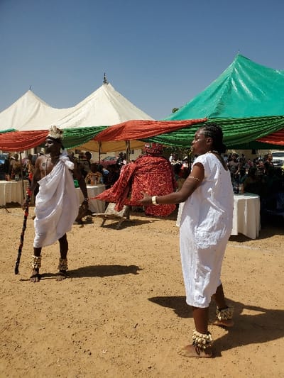 The scene depicts an outdoor gathering in Bar Gada, Nigeria, under a clear blue sky with bright sunlight. The foreground features two barefoot individuals on reddish-brown, sandy ground. On the left, an adult male with dark skin wears a white loincloth, an ornate light-colored headdress, and light-colored anklets. He holds a multi-banded staff vertically in his left hand and faces right. On the right, an adult female with dark skin wears a short-sleeved white tunic dress, light-colored anklets, and wrist bracelets. She extends both arms forward, manipulating or presenting a large, textured red fabric. In the background, multiple white and green peaked tents provide canopy cover, with additional red and green fabric drapes strung horizontally. Numerous attendees are visible under the tents, some seated at white-clothed tables. Shadows cast by the foreground figures are distinct on the ground.