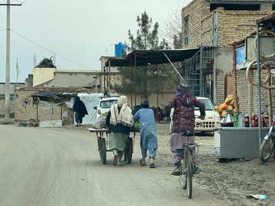 This image depicts an unpaved street scene in Nakhonay, Afghanistan, under an overcast sky. In the foreground, three individuals move away from the viewer. On the left, a person wearing a light green skirt/trousers and a white headscarf, and another person in light blue attire, are pushing a two-wheeled handcart loaded with dark bags and green leafy produce. To their right, a younger individual in a patterned padded jacket, brown trousers, and a beanie hat rides a dark-colored bicycle.

On the immediate right, a makeshift street vendor stall features a grey metal surface holding two kettles (one silver, one black), several plastic bottles (green, clear, dark), and branded soda bottles (Coca-Cola, Fanta). Behind this stall, yellow sacks are stacked, and a motorcycle is partially visible on the far right.

The midground and background consist of various structures. On the left, a utility pole stands near a low-rise building with exposed rebar, adjacent to a crude shelter constructed from various materials, including corrugated metal and tarpaulin. A person fully covered in a dark garment walks away from the viewer near this shelter, and a white vehicle (possibly an SUV or van) is parked, partially obscured. A blue tarp covers objects on the vehicle's roof. Further left, a white flag is visible on a pole in the distance.

On the right side of the street, a multi-story brick building shows signs of wear and construction, featuring an external metal spiral staircase. Behind the buildings, bare trees are visible, and an elevated blue cylindrical water tank is situated above the rooftops. The ground is unpaved, displaying dirt and potential muddy tracks.