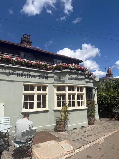 The image shows the exterior of "The Rising Sun," an independent beer house located in Epsom and Ewell, United Kingdom. The building is painted a pale green and features traditional windows with white frames. Flower boxes filled with pink and white flowers line the upper part of the building. The sign above the windows reads "THE RISING SUN" in large, dark lettering, with "INDEPENDENT BEER HOUSE" and "EST. 1871" also visible.

In the foreground, a person wearing a grey hoodie and blue shorts is sitting on a chair at a table outside the establishment. They are facing away from the camera, and their back is to the building. The scene appears to be set on a sunny day, indicated by the bright blue sky with white clouds and the strong shadows cast on the pavement. The pavement is paved with large, grey flagstones. A street runs alongside the pavement, with visible road markings. The overall atmosphere is relaxed and casual, suggesting a pleasant outdoor seating area at a pub.