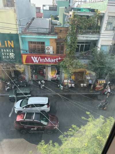 The image provides an overhead, high-angle view of a vibrant urban street scene, likely in Quy Nhon Ward, Vietnam, given the context. The perspective looks down onto the street, which runs diagonally from the bottom-left to the top-right of the frame, with buildings primarily lining the left side.
Several multi-story buildings are visible, showcasing a mix of residential and commercial establishments. One building is particularly green, covered in abundant foliage and flowering plants. At street level, a prominent red and yellow sign for "WinMart+" is clearly visible. Directly below it, a green sign identifies "LIN DANCE STUDIO" with the address "403A NGUYỄN HUỆ" and phone number "0766 72 73 57". Other businesses include small cafes or eateries with outdoor seating and a food cart marked "Bánh mì". Smaller signs with text like "20" and "SIÊU SỐC" are also present near WinMart+.
The street is bustling with activity. Numerous motorbikes, some carrying passengers, are either parked or in motion. Two cars are prominently parked near the right side of the street: a white SUV and a dark red SUV. The red SUV has "MẮT HÍTNH" written on its side. Several people are visible, either riding motorbikes, standing, or moving on the sidewalks. Overhead power lines crisscross the scene, a common feature in many Southeast Asian cities. The bright sunlight and clear shadows indicate it is daytime with fair weather. Lush green trees are interspersed throughout the urban landscape, adding natural elements to the concrete environment.