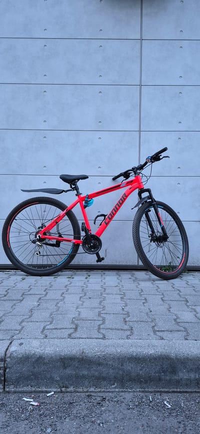 A bright red mountain bike is parked on a sidewalk in front of a concrete wall. The bike has black tires with red rims and a black seat. The brand name "CONNOR" is visible in large, white letters on the bike's frame. The concrete wall behind the bike is made of large rectangular panels with circular indentations. The sidewalk is paved with interlocking gray bricks, and a concrete curb is visible in the foreground, littered with cigarette butts. The scene appears to be in an urban environment, likely in the city of Ussurijsk, Russia. No people are present in the image.