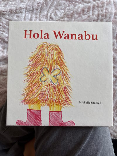 A book cover titled "Hola Wanabu" by Michelle Shubich is in the center of the image. The cover features a drawing of a person's head with yellow and red hair and a blue hair clip and red boots. The book is resting on a fluffy grey blanket, next to a person wearing grey denim pants. The location is not discernible from the image.