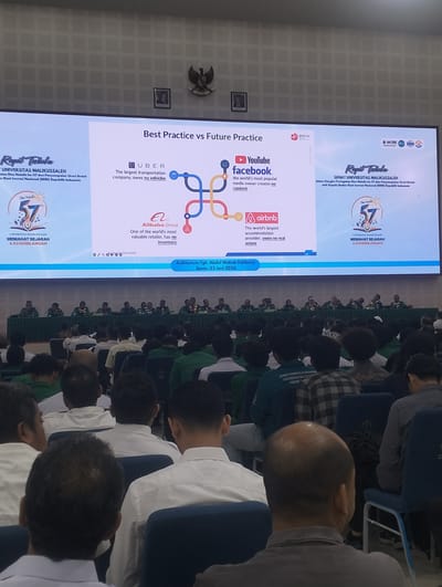 This image shows a large screen displaying a presentation titled "Best Practice vs Future Practice" in what appears to be an auditorium. The screen content includes logos and descriptions of companies like Uber, YouTube, and Airbnb, along with text related to "UNIVERSITAS MALIKUSSALEH" and "MEMAHAT SEJARAH & KEGEMILANGAN". On the left side of the screen, there's a graphic with a "Dies Natalis 57" badge. At the bottom of the screen, it indicates "Auditorium Tgk. Abdul Wahab Dahlawy, Senin, 22 Juni 2026". The foreground shows an audience of people seated, facing the screen, with some of them wearing uniforms. The overall scene suggests an academic or formal event taking place in Indonesia, specifically in the Auditorium Tgk. Abdul Wahab Dahlawy, which is located in Reuleut Timu, Indonesia.