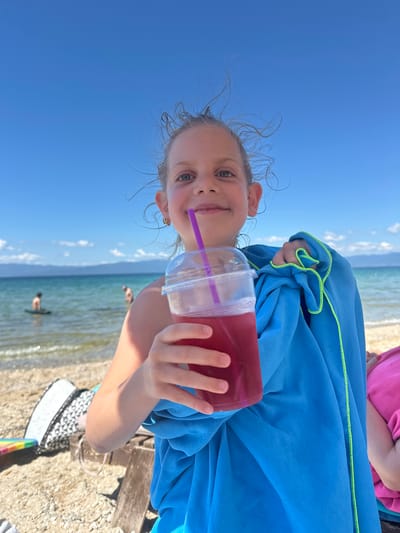 An eye-level, medium shot captures a young girl on a beach in Ofrynio Beach, Greece, holding a cup with a purple straw and a blue towel. The girl has blonde hair and is smiling at the camera. The blue towel is draped around her and held by her left hand. The drink in the cup is a deep pink or red color. The background shows a sandy beach with some beach chairs and bags, the turquoise sea, and distant hills under a clear blue sky. Two people are visible in the water in the background. The image is taken on a sunny day.