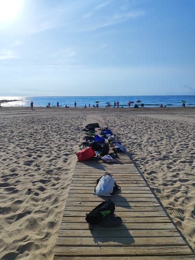 The image depicts a wooden boardwalk on a sandy beach in El Campello, Spain, with the Mediterranean Sea in the background. The boardwalk is lined with various bags and shoes, suggesting people have left their belongings to enjoy the water or activities. Several individuals are visible in the distance on the beach and in the sea, some under umbrellas. The sky is clear blue with some wispy clouds, and the sun is shining brightly, casting shadows on the sand and boardwalk. The scene conveys a typical summer day at a coastal resort.