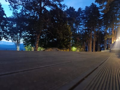 The image depicts a low-angle view of a wooden deck with a visible wood grain texture. In the background, a line of tall pine trees stands against a dusky blue sky. The trees are silhouetted, with some branches extending into the foreground. Artificial lights are illuminating parts of the trees and the ground behind the deck, creating distinct shadows on the deck's surface. The lighting suggests it is dusk or nighttime. In the distance, a faint outline of mountains and a body of water can be observed. The scene appears to be an outdoor area, possibly a park or a viewpoint overlooking a landscape. Based on the provided context, this location is the "Hisar" area in Kyustendil, Bulgaria, known for its natural beauty and recreational spaces. There are no people visible in the image.