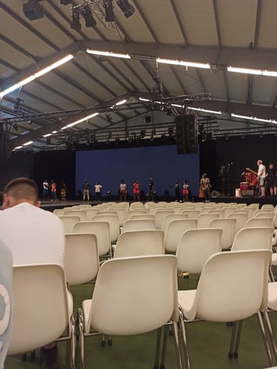 The image depicts an indoor event taking place in what appears to be an auditorium or large hall, possibly in Warffum, Netherlands. Rows of empty, modern, white plastic chairs are arranged on a green carpeted floor, facing a stage area. The chairs are neatly organized in multiple rows, suggesting a seated audience capacity.
On the stage, a performance or presentation is in progress. Several individuals are visible, some standing and others potentially seated. One person is playing a guitar, and another is standing near a drum set, indicating a musical performance. The stage is set against a dark backdrop, illuminated by stage lighting and overhead fluorescent lights that run along the ceiling structure. The ceiling itself is made of a light-colored, possibly fabric or tent-like material, with visible supporting beams and rigging for lighting and sound equipment.
The lighting suggests it is likely evening or an indoor event where external light is not a factor. The overall atmosphere seems to be one of anticipation or a mid-event calm, with the audience seats mostly empty, yet people are actively engaged on the stage. The scene is captured from a slightly elevated angle, looking down towards the stage and across the rows of chairs. There is no visible text in the image.