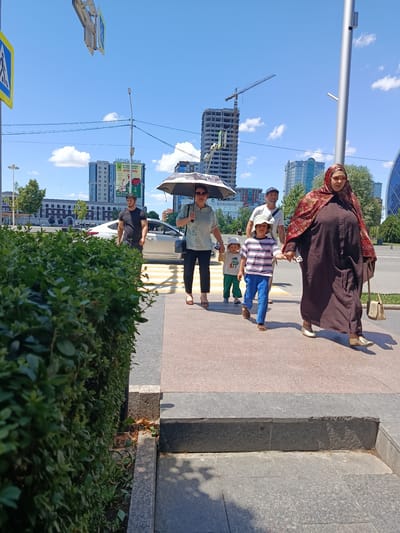 A group of six people are walking on a sidewalk next to a street in Grozny, Russia, on a sunny day. The group includes three adults and three children. Two of the children are walking in front, one of whom is being held by the hand by an adult. The adults appear to be walking behind them. The group is walking on a sidewalk with a pedestrian crossing in the background. In the background, there are several buildings, including a tall, unfinished building with a crane, and a street with cars. The scene is captured from a slightly low angle, with a lush green hedge in the foreground on the left side of the image.