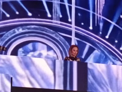 A performer, possibly a DJ, stands behind a barrier on stage in a venue that is likely an event hall, performance arena, or concert space located in San Bartolomé de Tirajana, Spain. The performer is seen from the chest up, with headphones around their neck. The backdrop features a visual display of concentric arcs with a neon or illuminated effect, punctuated by vertical streaks of light. The barrier is textured with a white and blue design. The lighting is generally blue-toned.