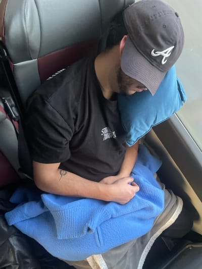A male individual is observed sleeping or resting in a vehicle seat, consistent with public transportation, within the inferred context of Leiria, Portugal. He is positioned with his head resting against a light blue rectangular pillow placed against a window. The subject wears a dark baseball cap with a white embroidered "A" logo, obscuring his upper face, and a dark short-sleeved t-shirt with a white graphic text, "STREET TRUTH," visible on the left chest area. A stylized tattoo, resembling a circle with a downward line, is visible on his right forearm. His hands are clasped together on his lap, which is partially covered by a light blue fleece garment. The seat features grey and maroon upholstery, with a seatbelt mechanism visible on the left side of the frame. No specific external landmarks identifying the precise location within Leiria are discernible.