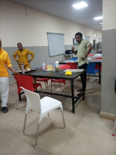 A brightly lit room features several people, tables, and chairs. Two women stand to the left, one wearing yellow and white clothing, the other wearing yellow. A man in a green shirt and black pants stands next to a table, gesturing. A whiteboard hangs on the wall in the background. The tables have red, white, and blue chairs around them. Food and drinks are arranged on the tables.