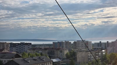 This image presents an elevated view of a residential area in Lensk, Russia. The scene depicts a quiet urban landscape dominated by apartment buildings, with a distinct layer of mist or fog in the distance.

In the foreground, a prominent dark diagonal wire or cable stretches from the upper right corner to the lower left. Below this, the dark roofs of several older, low-rise residential buildings are visible, interspersed with green trees.

The mid-ground is occupied by numerous multi-story apartment blocks. These buildings exhibit a range of architectural styles, including older, seemingly Soviet-era structures with uniform windows and weathered facades, and newer or renovated buildings featuring varied color palettes like black, white, blue, and orange panels. Lush green foliage from trees and bushes is scattered throughout this urban fabric.

In the background, a distinct, horizontal band of low-lying mist or fog blankets the landscape, creating a hazy horizon line. Above and beyond this mist, the faint, dark silhouettes of distant hills or forested areas can be discerned.

The sky is largely covered by stratocumulus clouds, which are broken enough to allow diffused sunlight to illuminate the scene, casting soft light and shadows. The presence of mist suggests either early morning or late evening, though the overall brightness leans towards a morning setting. No individuals or specific activities are visible, lending a serene atmosphere to the urban panorama. There is no discernible text within the image.