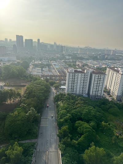 The image displays a high-angle, wide shot of a hazy urban landscape in Subang Jaya City Council, Malaysia, likely captured during early morning or late afternoon due to the soft, diffused light and visible haze.
In the foreground, a road curves into the frame, bordered by dense green foliage and trees. The road surface appears wet or reflective, with yellow pedestrian crossings marked. A red vehicle is seen traveling on the road, and further down, a dark-colored utility vehicle is parked. A group of people are visible standing along the side of the road, seemingly gathered on a bridge or elevated walkway.
Beyond the immediate roadside greenery, a suburban area unfolds with rows of terraced houses, their roofs predominantly orange and brown, interspersed with parked cars. Several multi-story apartment buildings, white with brown roofs, are prominent features in the mid-ground.
The background reveals a dense urban skyline characterized by numerous high-rise buildings of varying heights and architectural styles, receding into the atmospheric haze. The overall impression is of a sprawling city with a mix of residential, commercial, and natural elements, under a soft, possibly polluted, sky.