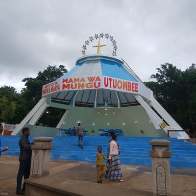 The image shows the Grotto of the Blessed Virgin Mary in Moshi, Tanzania, with a distinctive conical building topped with a gold cross surrounded by a ring of stars. A banner in red text proclaims "MAMA WA MUNGU UTUOMBEE" and "GROTTO YA BIKRA MARIA." Blue stairs lead up to the entrance of the building, and blue chairs are visible inside. Several people are on the steps, including two women and a child wearing patterned outfits, and a man wearing a gray shirt and dark pants. A low, textured stone pillar stands on the side of the stairs. The sky is overcast, and trees are visible in the background.