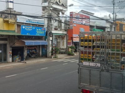 The image shows a street scene in B'Lao, Vietnam, on an overcast day. In the foreground, the rear of a metal truck is visible, laden with colorful plastic crates, primarily yellow, green, and red. A license plate on the truck reads "50H-687.78". The truck is positioned on the right side of a paved road with white lane markings. A pedestrian crossing is visible further down the road.
On the left side of the road, a row of buildings lines the street. The closest building has a green awning and a blue sign with Vietnamese text and an image of a bird. Further back, a yellow building with a water tank on its roof is visible. Above the blue sign, another larger banner with Vietnamese text is partially obscured by wires.
Across the street, several businesses are identifiable by their signs. One prominent orange building features a red sign with Vietnamese text, including a phone number, and the word "Tiên". Another sign above it is partially visible, displaying Vietnamese text and what appears to be an advertisement. Further down the street, other buildings with various signs and balconies are present, along with numerous electrical wires strung across the scene. Two motorcycles are parked on the sidewalk in front of one of the buildings. Vegetation, including small trees and potted plants, can be seen near some of the structures. The overall impression is a busy urban environment with typical street infrastructure.
