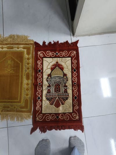 Two Islamic prayer rugs are displayed on a white tiled floor. The rug on the left is a golden-brown color with a woven palm tree design. The rug on the right is red with a central cream-colored panel depicting a mosque, possibly the Taj Mahal, with intricate golden and red detailing. Both rugs have fringed edges. A person's feet, wearing grey socks and a blue pant leg, are visible in the lower portion of the image. The background consists of white tiled flooring and a portion of a light-colored wall or structure. The scene appears to be indoors.