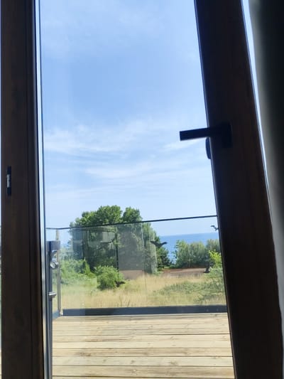 The image depicts a view through an open doorway of a room, looking out onto a deck and a coastal landscape. The doorway has wooden frames and a dark-colored handle on the right side. The deck is made of light-colored wooden planks laid horizontally. Beyond the deck, there is a glass railing, and beyond that, a grassy area with scattered wildflowers. Further in the background, there are lush green trees, and in the distance, a body of water, likely the sea, stretches to the horizon under a bright blue sky with scattered white clouds. On the glass railing, two dark bird-shaped decals are visible, one on the left and one on the right, appearing to be in flight. The location is specified as Lozenets, Bulgaria, a coastal town known for its beaches and natural beauty, which aligns with the scene presented. The scene is one of a serene outdoor setting, likely a private balcony or patio area of a residence or accommodation.