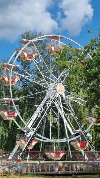 A white Ferris wheel with red and gold gondolas stands outdoors, surrounded by lush green trees under a partly cloudy blue sky. The Ferris wheel has a central medallion with the words "FERRIS WHEEL" inscribed on it. The base of the Ferris wheel is decorated with colorful imagery and the words "FERRIS WHEEL" in large, ornate lettering. The scene is set in the city of Smila, Ukraine.