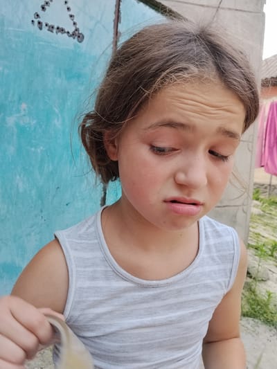 A close-up shot features a young girl with brown hair pulled back in a braid, her face turned downwards and her eyes closed. She wears a light grey striped tank top. Her expression appears distressed, with furrowed brows and a slightly downturned mouth. The background is a textured, faded turquoise wall with some dark markings, possibly bullet holes, forming a triangular shape on the upper left. To the right, a hint of a concrete structure and some green foliage are visible, along with a blurred pink garment hanging in the distance. The lighting suggests it is daytime, with the overall scene conveying a sense of outdoor activity, possibly in a rural or less developed area. The girl's hands are visible in the lower left corner, holding a light-colored object.