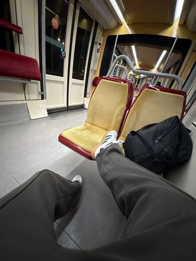 Tan and maroon train seats are occupied by a black backpack and a person wearing gray sweatpants and sneakers with white uppers and tan soles. One foot rests on the seat in front, while the other is on the floor. Windows line the left side, with a glimpse of red seating further down.