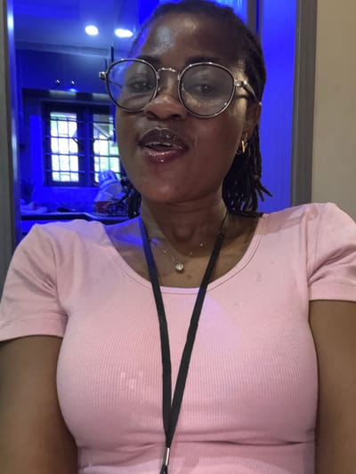 A woman with braided hair and glasses is pictured from the chest up. She is wearing a pink, ribbed t-shirt with a black lanyard around her neck. Her mouth is slightly open and she appears to be speaking. The background is out of focus and shows a bright blue light and what appears to be a window with grids. The image is taken indoors, possibly in a public building or a space with decorative lighting. The overall impression is a candid shot, possibly a selfie, given the angle and close-up framing of the subject. The scene is brightly lit, with the blue light creating a vibrant and somewhat abstract backdrop.