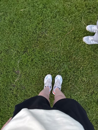 Two pairs of feet, one pair on the left wearing white athletic shoes with blue accents and black shorts, and another pair on the right wearing light grey athletic shoes with white laces, are standing on a grassy area. The grass appears to be outdoors and possibly in a park or yard. There are no visible buildings or other structures in the immediate vicinity. The scene does not provide any specific clues to determine the city of Otchor, Russia, or any other urban location.