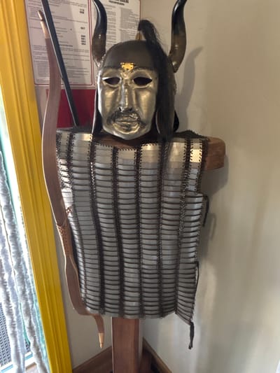 An ornate, metallic mask with a mustache and beard is worn by a bust. The mask has two prominent horns, one on each side, and dark hair cascading from the top and sides. The bust is clad in a sleeveless shirt made of chainmail, with rows of silver-colored metal plates linked together by dark fastenings. The chainmail extends down to cover the torso. The bust is mounted on a wooden display stand. To the left of the bust, a wooden bow is visible, leaning against a yellow surface that appears to be a curtain or partition. Behind the bust, a wall is visible, and to the left, a window covered with a white, textured curtain. The overall scene suggests a museum or exhibition display of historical armor.