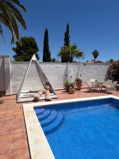 This is a photograph of a backyard swimming pool area in Mont-roig del Camp, Spain. The pool is filled with bright blue water and features tiled steps. The surrounding patio is paved with terracotta tiles. To the left of the pool, there is a white triangular cabana with a light-colored mesh covering and a large, tan teddy bear sitting on a cushion inside. In the background, there is a white textured wall, potted plants, a dining table with chairs, and several trees, including palm trees and tall, slender evergreens. The sky is clear and blue, indicating a sunny day.