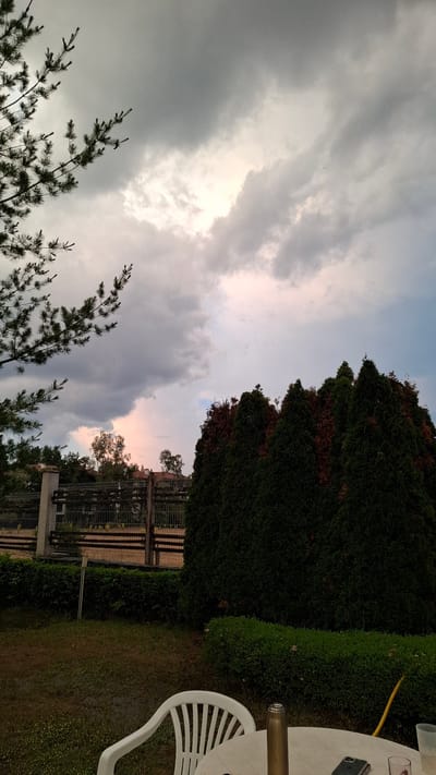 This is a low-angle, vertical photograph of an outdoor scene, likely a backyard or garden, under a dramatic sky. The main subjects are the sky filled with heavy, grey clouds that are beginning to break, allowing soft, pink and orange light to filter through, suggesting either dawn or dusk. In the foreground, silhouetted pine branches reach up towards the sky. To the right, a dense, dark green hedge or tall shrub forms a natural wall, with a portion of a white plastic table and chair visible, along with a glass and a metallic object that could be a bottle. A yellow hose is draped over the table. In the middle ground, there are glimpses of a fence and some trees, with some areas of the hedge showing signs of browning. The overall mood is serene, with a sense of approaching change in the weather, possibly a storm or the end of one. There is no visible text. The location is identified as Razlog, Bulgaria.