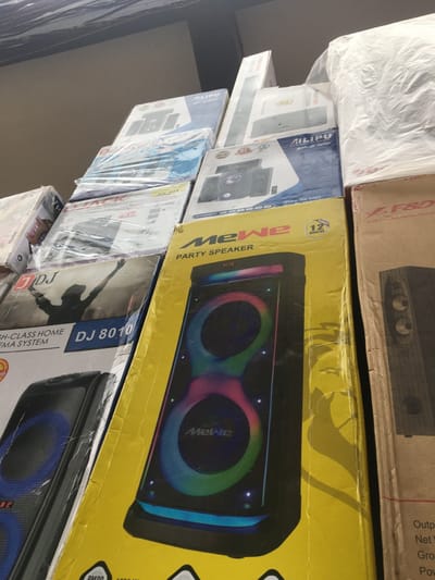 The image, captured in Ojo, Nigeria, depicts a retail environment featuring a stack of boxed electronic goods, primarily audio equipment. In the foreground, a prominent yellow cardboard box advertises a "MeWe PARTY SPEAKER," displaying an image of a black, vertical speaker with two circular, multi-colored LED illuminated woofers and a glowing base. Text "12 MONTHS" is visible on this box. To the immediate left, a box for a "DJ 8010 HIGH-CLASS HOME CINEMA SYSTEM" shows a black silhouette of a person with arms raised. Further boxes are stacked behind and around these, displaying brands such as AILIPU and F8D, and imaging other speakers, sound bars, and home theatre systems. Many boxes are individually wrapped in clear plastic. The background shows a light-colored wall or ceiling junction. No individuals are visible in the scene.