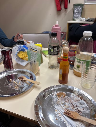 The image depicts an indoor scene on a light-colored table in West Lancashire, United Kingdom, following a casual meal or gathering. In the foreground, two silver oval paper plates are visible; both contain dark food residue, consistent with dessert or cake remnants, and each has a light-colored wooden fork resting on it.

On the table, various items are distributed:
- To the left, a red aluminum beverage can stands next to a plate.
- A partially open clear plastic bag contains yellow tortilla chips.
- Adjacent to the chips, a clear plastic pouch holds a light-colored dry food product.
- Beverage containers include an empty clear plastic bottle with a black cap (identified as Coca-Cola Zero Sugar), a pink plastic water bottle with a straw-top, and a clear 1-liter plastic bottle of apple juice (featuring a green apple logo).
- Condiments consist of an amber-colored glass bottle of hot sauce with a wooden stopper and a partially obscured clear plastic bottle of Heinz Tomato Ketchup.
- An empty, clear ribbed glass tumbler is positioned on the right.

Regarding people, the left arm of an individual is visible, clad in a dark blue long-sleeved garment, with blue-painted fingernails, holding a small, clear object. The arm and shoulder of a second individual, wearing dark clothing, are partially visible on the far right of the frame.

The background shows a plain, light-colored wall. A light-colored chair backrest is visible centrally. To the right, a wooden shelf or cabinet is present, above which a green food waste bin, labeled "Food Waste" with corresponding icons, is mounted on the wall. A red item, possibly clothing, hangs on the wall further left in the background.