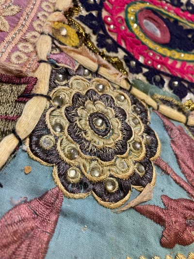 This is a close-up, overhead shot of a heavily embroidered textile. The embroidery is intricate and features a variety of colors and textures. In the foreground, a circular, floral-like motif is dominant, rendered in dark purple and gold thread. This central motif is adorned with numerous small, clear beads. Surrounding this, other embroidered elements are visible, including pink and purple leaf-like shapes, as well as strands of beige yarn tied with dark thread. Further back, portions of a larger, more abstract design are present, incorporating bright pink, yellow, and dark blue fabric, interspersed with gold thread and more small beads. The background textile is a light blue-green. There are no people or discernible environmental features visible beyond the textile itself. The image does not contain any information to suggest a location in Sabadell, Spain.