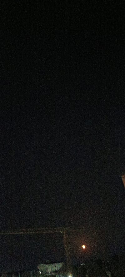 This image captures a nighttime scene in Fergana City, Uzbekistan. The sky is a deep, dark black, speckled with a significant amount of digital noise, obscuring any stars or celestial bodies. In the lower portion of the image, a dimly lit landscape is visible. There is a faint horizontal structure that appears to be a building or an elevated walkway stretching across the scene. To the right of center, a bright, circular light source glows, possibly a streetlamp or a distant building light, casting a warm, orange hue. Below this light, and also in the lower part of the image, are indistinct shapes that suggest buildings or structures, some of which are dimly illuminated. The overall impression is a quiet, dark urban environment at night, with limited visibility and a focus on the sparse artificial lights that punctuate the darkness. The grainy texture of the image suggests it was taken with a low-light setting or in challenging lighting conditions.