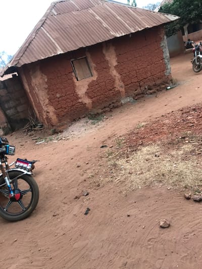 The scene in Ogodo, Nigeria, displays an unpaved, reddish-brown dirt surface occupying the foreground and midground, marked with tire tracks and scattered small rocks and dry vegetation.
A single-story structure constructed of reddish-brown, cracked mud or packed earth walls with an uneven texture dominates the midground. It features a weathered, corrugated metal roof. On the visible facade, a single rectangular window with closed wooden shutters is present. An incomplete concrete block wall section abuts the left side of this building.
In the foreground on the left, the front wheel and partial body of a black motorcycle are visible. It has a chrome front fender, a black tire with a multi-spoke silver rim, and a red circular accent on the hub. A blue rectangular plate with white lettering, partially reading "ONOKING," is attached to the front.
In the background on the right, a second motorcycle, appearing white or silver, is partially visible, parked or in motion near a lighter-colored building partially obscured by green foliage. A faint human figure is discernible near this background motorcycle, but specific details are not resolvable.
