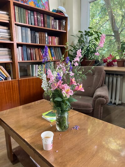 A still life photograph captures the interior of a room, possibly a library or study, in Pravets, Bulgaria. The foreground is dominated by a wooden table with a slightly worn, polished surface. On the table, a glass vase holds a vibrant bouquet of pink and purple flowers, a few small dried flower petals are scattered nearby. To the right of the vase is a paper coffee cup, partially filled with a dark liquid, showing a portion of its white and pink patterned exterior. Behind the table, a plush brown armchair is situated near a window. Beyond the armchair, a radiator is visible. A tall wooden bookshelf filled with numerous books stands to the left of the armchair and table. The books are of various sizes and colors, with some spines displaying Cyrillic text. Above the books, a colorful painting hangs on the wall. The window offers a view of lush green trees outside, suggesting an outdoor environment with abundant foliage. The overall impression is one of a tranquil and lived-in space.