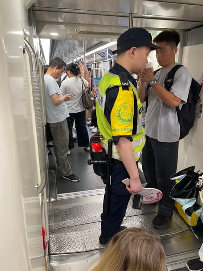 The image depicts the interior of a crowded subway car in Shenzhen, China, not Hong Kong. A transit police officer wearing a bright yellow safety vest over a dark blue uniform stands in the foreground. The vest has patches with Chinese text and emblems. The officer has a baton clipped to their belt and is holding a white circular sign with red markings. To the officer's right, a young man with short hair and white earbuds is looking down, his hands clasped together. He wears a grey t-shirt and black pants, with a black backpack slung over one shoulder. Behind the officer, another man in a light blue t-shirt and grey cargo pants is looking at his phone. Several other passengers are visible in the background, some standing and others seated. The subway car interior is utilitarian, with metal grab bars and a diamond-plated floor in the entryway. A red fire extinguisher is mounted on the wall to the left of the officer.
