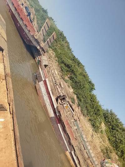 This image, taken in Ibi, Nigeria, depicts a waterfront scene with a strong diagonal composition. A body of murky, brown-tinged water occupies the lower-left portion of the frame. Along its right edge, a steep, earthen bank rises, supporting a line of at least six long, narrow wooden boats moored parallel to the shore. One prominent boat in the foreground features the name "YUSUF" painted in white on its side, alongside faded red and white stripes. Other boats display weathered, unpainted wood or remnants of faded paint. The bank transitions into a densely vegetated hillside covered with green bushes and small trees, which extends towards the upper-right. The sky above the vegetation is clear and light blue. In the bottom right corner of the frame, a partial view of an individual's head and shoulder, adorned with multi-patterned fabric, is visible. No direct actions or interactions are depicted, with the boats appearing stationary.